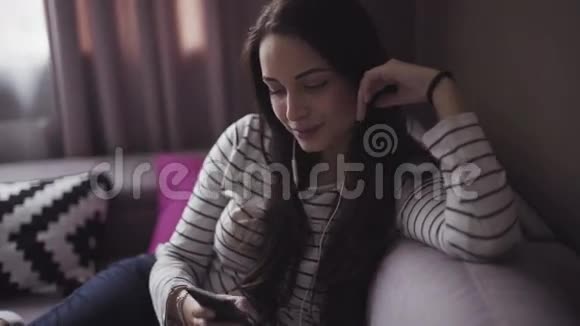 年轻的女人坐在客厅的沙发上女孩通过耳机听她的移动设备的音乐视频的预览图