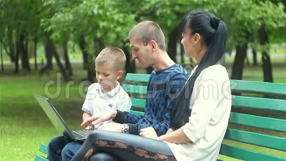 爸爸妈妈和儿子的家人坐在公园的长凳上看着一台笔记本电脑视频的预览图