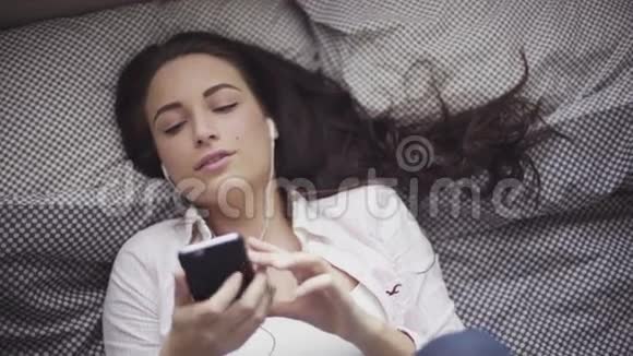 迷人的白种人女孩躺在床上听着她的移动设备耳机插入的音乐视频的预览图