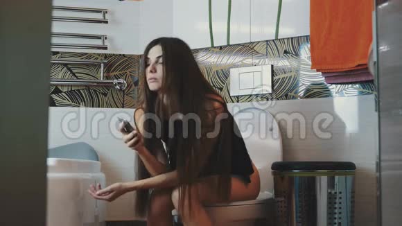 布鲁内特年轻女孩坐在马桶上手里拿着智能手机洗手间视频的预览图
