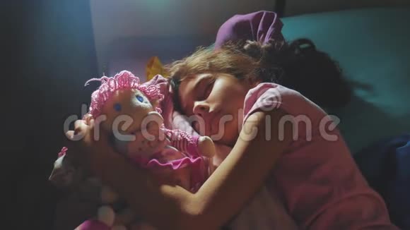 可爱的小女孩睡在沙发床上拥抱她的娃娃晚上睡在床上的黑发小女孩视频的预览图