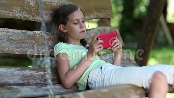 带红色智能手机的女孩坐在花园里的秋千凳上听音乐视频的预览图