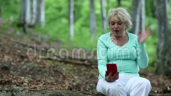 女人坐在森林中一棵倒下的树上通过她的智能手机交流视频的预览图