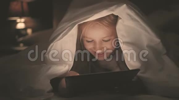 在黑暗的光线下躺在床上的少女在社交网络上玩平板电脑特写小女孩看视频的画面视频的预览图
