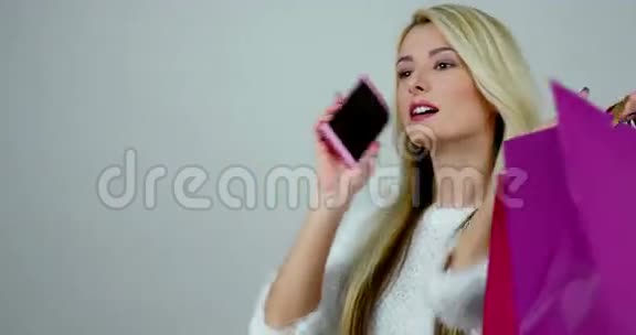 一个年轻兴奋的金发女人在手机上交谈的肖像持有五颜六色的购物包视频的预览图