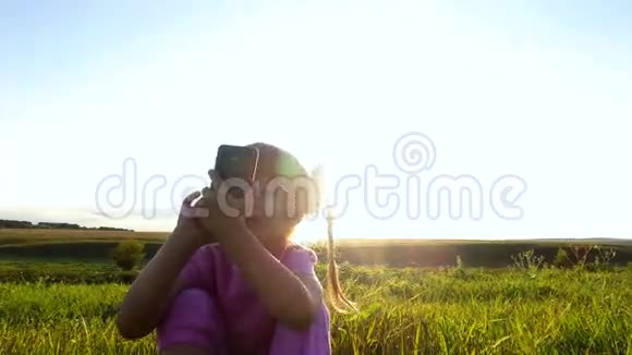 热情的小女孩在智能手机上玩戴眼镜的女孩坐在绿色的草地上视频的预览图