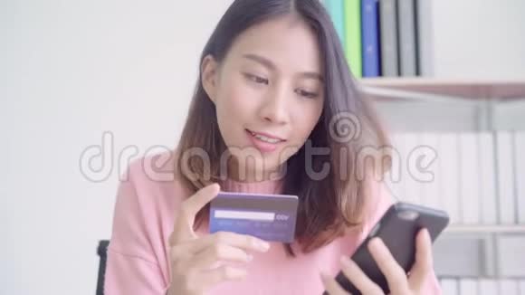 亚洲女性使用智能手机通过信用卡购买网上购物而穿毛衣坐在家里客厅的桌子上视频的预览图