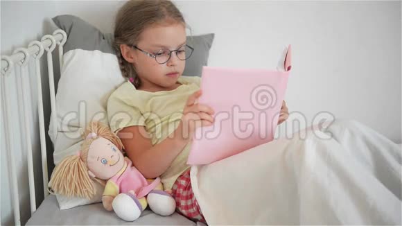 戴眼镜的小女孩躺在床上看书她旁边坐着一个洋娃娃视频的预览图