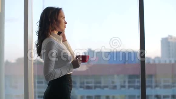 欧洲现代年轻漂亮的女商人站在全景窗边喝咖啡发信息视频的预览图