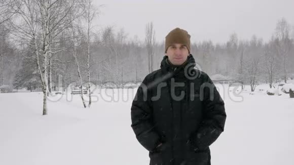 年轻人看着镜头在冬林中微笑大雪纷飞一个穿着深色夹克戴着暖和帽子的男人视频的预览图