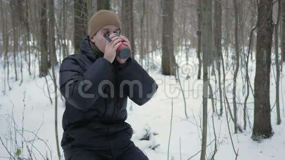 一个年轻人在冬天的森林里用热水瓶喝热饮料那人在雪地里环顾四周在树上视频的预览图