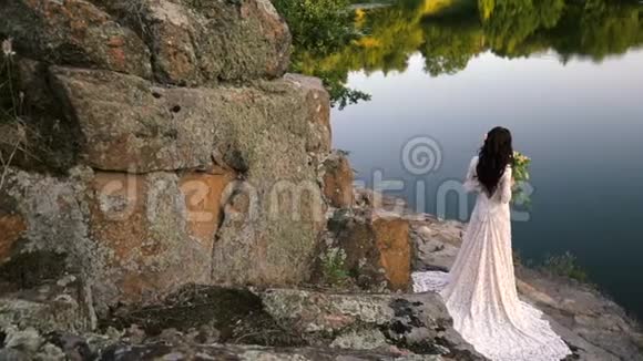 年轻的新娘站在岩石河岸日落视频的预览图
