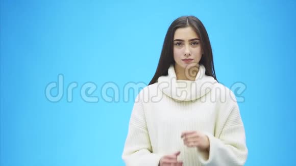 美丽的年轻女孩站在蓝色的背景上在这段时间里她穿着一件白色毛衣很长的黑色视频的预览图