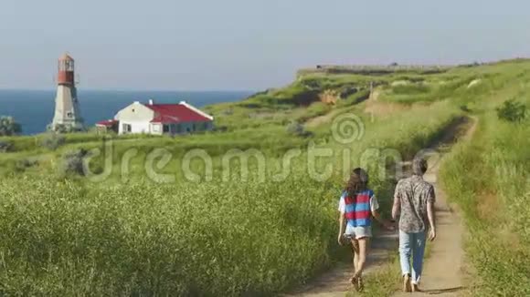 年轻夫妇在阳光明媚的天气走在海岸线附近背景是浪漫的灯塔背面视频的预览图