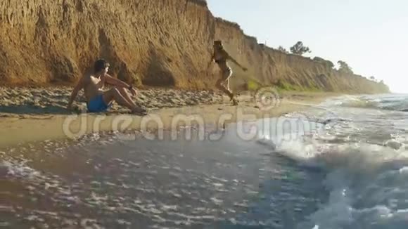 快乐的一对夫妇在沙滩上有高高的砂岩悬崖年轻活泼的女孩在英俊的坐着男人身边嬉戏视频的预览图