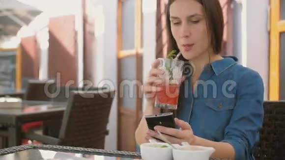 布鲁内特女孩使用智能手机而坐在外面的咖啡馆喝和享受凉爽的饮料从吸管4k视频的预览图