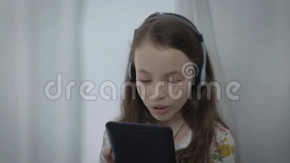 漂亮的小女孩带着耳机在平板电脑上观看音乐视频并唱歌视频的预览图