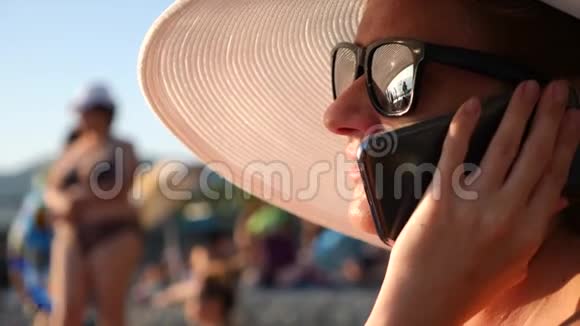 4K女人戴着白色宽边帽子和太阳镜在海滩上用她的手机慢动作视频的预览图
