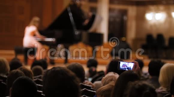 在演奏钢琴女孩时的音乐厅观众人们在智能手机上拍摄演出音乐歌剧视频的预览图