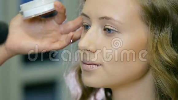 专业化妆师在金色卷发和蓝眼睛模型皮肤上涂上色调霜刮脸视频的预览图