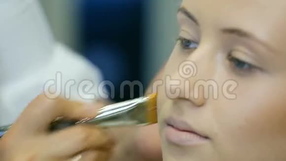 专业化妆师在金色卷发和蓝眼睛模型皮肤上涂上色调霜刮脸视频的预览图