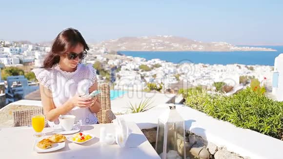 白种人女孩早餐与智能手机在户外咖啡馆令人惊叹的景观Mykonos镇视频的预览图