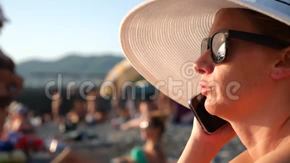 4K女人戴着白色宽边帽子和太阳镜在海滩上用她的手机慢动作视频的预览图