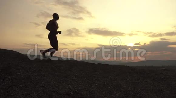 在山顶日落时慢跑的非裔美国人运动员的侧影多云的天空背景视频的预览图