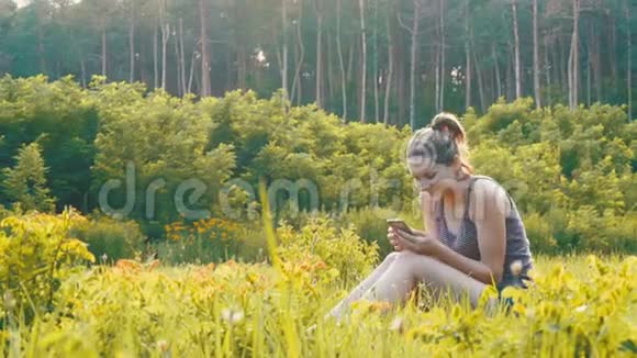 快乐的年轻女子坐在绿色的草坪上在夕阳的背景下在风景名胜区使用智能手机视频的预览图