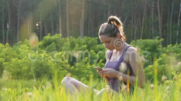 快乐的年轻女子坐在绿色的草坪上在夕阳的背景下在风景名胜区使用智能手机视频的预览图