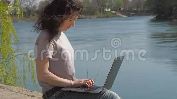 一个女孩正拿着笔记本电脑坐在河岸上一个春天阳光明媚的日子带着笔记本电脑在水里的年轻女子视频的预览图