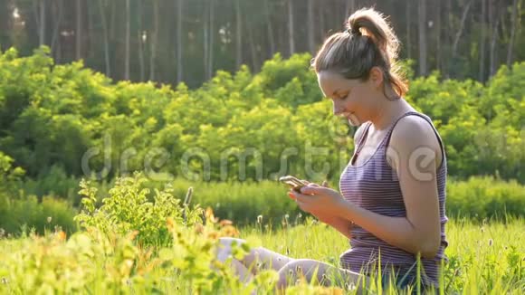 快乐的女孩坐在绿色的草坪上在日落时使用智能手机视频的预览图