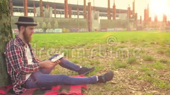 年轻的嬉皮士在公园里的一棵树旁边放着一本放松阅读的书视频的预览图