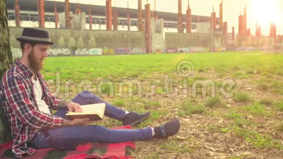 年轻的嬉皮士在公园里的一棵树旁边放着一本放松阅读的书视频的预览图