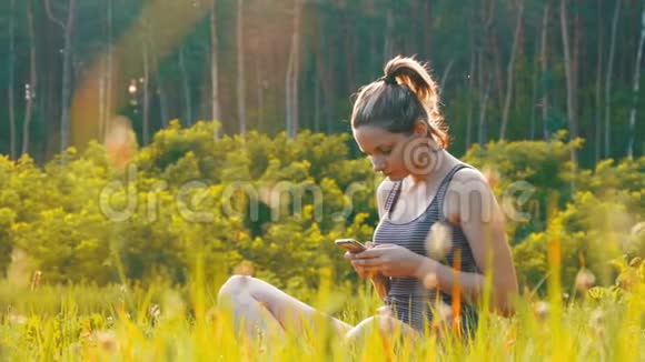 坐在绿色草坪上在日落背景下的风景区使用智能手机的快乐年轻女子视频的预览图