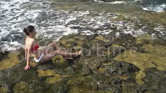 年轻漂亮的泳装女郎微笑着坐在海边的岩石上视频的预览图