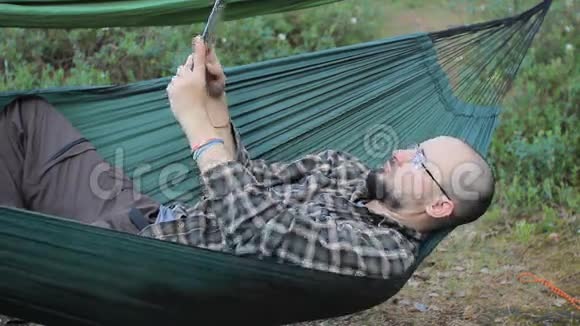 一个穿吊床的男人在平板电脑上自拍一个留着胡子戴眼镜的人视频的预览图