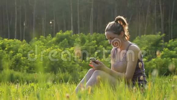 坐在绿色草坪上在日落背景下的风景区使用智能手机的快乐年轻女子视频的预览图