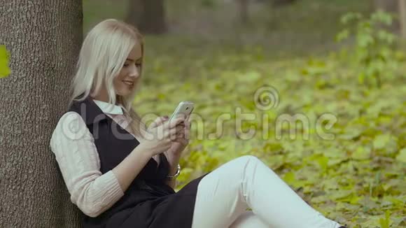 秋天公园里一个美丽的女孩坐在树下手里拿着手机微笑着看着留言视频的预览图