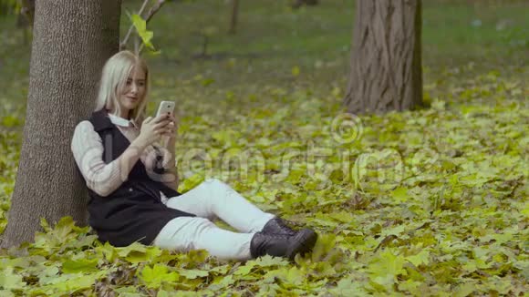秋天公园里一个美丽的女孩坐在树下手里拿着手机微笑着看着留言视频的预览图