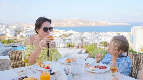 一家人在室外咖啡馆吃早餐在Mykonos镇有令人惊叹的景色可爱的女孩和妈妈喝着新鲜果汁视频的预览图