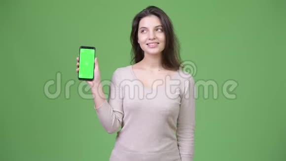 年轻快乐的美女一边思考一边在绿色背景下显示手机视频的预览图