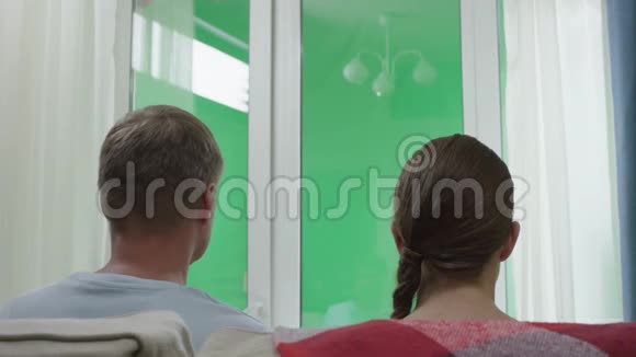 一对夫妇拥抱坐在沙发上望着室外透过窗户看到绿色的屏幕视频的预览图