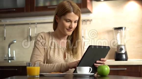 美丽的年轻迷人的女人吃早餐在厨房里浏览互联网视频的预览图