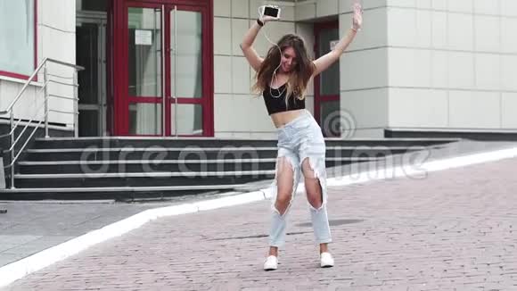 年轻女孩在街上跳舞在耳机上听音乐很有趣穿着破牛仔裤和背心的漂亮女孩视频的预览图