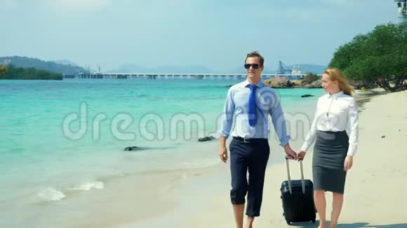 商人和女商人带着一个手提箱沿着岛上白色的沙滩散步视频的预览图