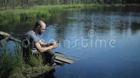 一个人坐在湖边的一个码头上摸着石碑背景上美丽的蓝色湖泊和森林视频的预览图