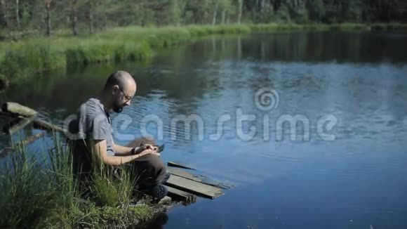 一个人坐在湖边的码头上玩平板游戏背景上美丽的蓝色湖泊和森林视频的预览图
