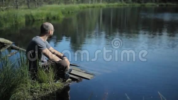 一个人坐在湖边的码头上触摸智能手表背景上美丽的蓝色湖泊和森林视频的预览图