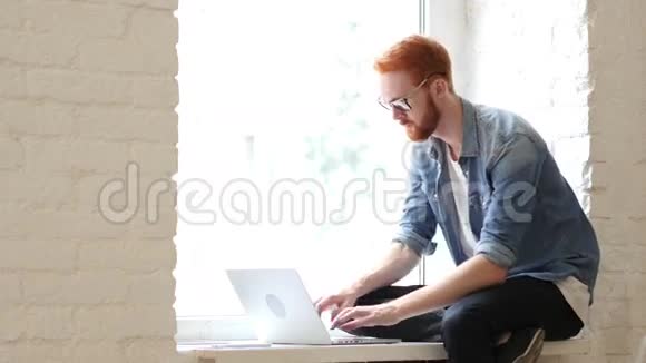 穿着笔记本电脑打字坐在窗前放松的男士视频的预览图